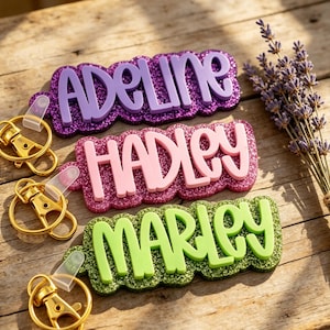 Könnte beinhalten: Drei personalisierte Schlüsselanhänger mit erhabener Schrift auf glitzernden Hintergründen. Der oberste Anhänger ist lila mit dem Namen ADELINE, der mittlere ist rosa mit HADLEY und der untere ist lindgrün mit MARLEY. Jeder hat einen goldenen Verschluss.