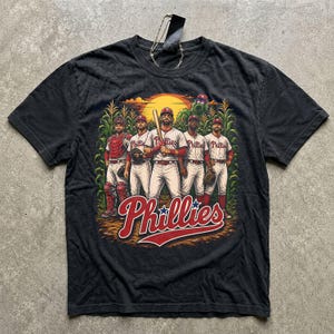 May include: A charcoal gray short-sleeve t-shirt featuring a graphic of five baseball players in white pinstripe uniforms standing in a cornfield. The text Phillies is printed in red script at the bottom with a sunset in the background.