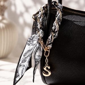 Könnte beinhalten: Eine schwarze, texturierte Handtasche mit einem dekorativen Riemenanhänger aus einer goldfarbenen Metallkette, die mit einem weißen Stoffband mit schwarzem botanischem Blattmuster verflochten ist, und einem hängenden Buchstaben-S-Anhänger.