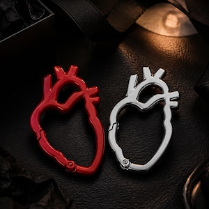 Könnte beinhalten: Zwei Karabiner in Form eines anatomischen Herzens, einer in glänzendem Rot und der andere in poliertem Silber. Jeder Karabiner verfügt über einen federbelasteten Verschlussmechanismus an der Basis, der die Form eines menschlichen Herzens hat.