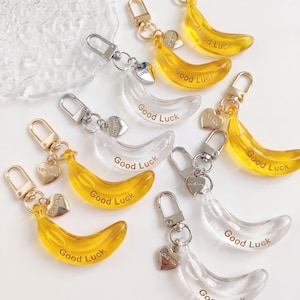 Könnte beinhalten: Eine Sammlung von bananenförmigen Schlüsselanhängern aus transparentem und gelbem Acryl. Jeder Anhänger hat einen goldfarbenen Karabinerhaken, ein kleines herzförmiges Plättchen mit der Gravur Love You und den Schriftzug Good Luck.