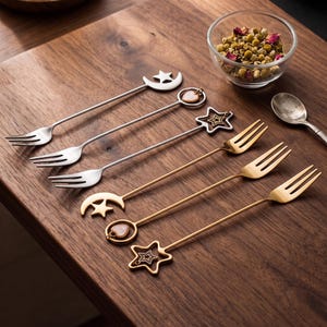 May include: A set of six long-handled metal dessert forks in silver and gold tones. The handles feature decorative charms including crescent moons, stars, and heart-shaped pendants. These slender forks are arranged in a diagonal row.