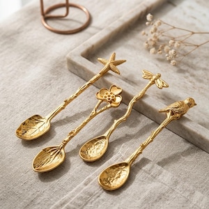 Peut inclure: Un ensemble de quatre cuillères en métal doré avec des bols texturés et des manches en forme de branche. Les manches sont surmontés de motifs décoratifs distincts : une étoile de mer, une fleur à quatre pétales, un insecte et un petit oiseau.