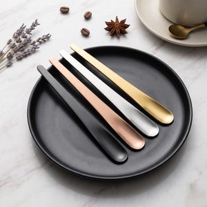 May include: A set of four long-handled spoons in black, copper, silver, and gold, arranged on a black plate. The spoons have a sleek, modern design, perfect for stirring coffee or tea. The background includes coffee beans and a star anise.