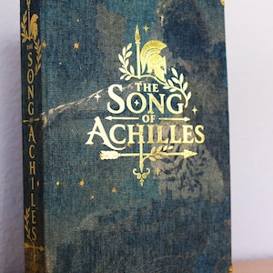 May include: A hardcover book titled The Song of Achilles by Madeline Miller with a dark blue textured fabric cover. The front and spine feature gold foil stamping of a helmet, arrows, and stars. Gold metal corner protectors are on the cover.