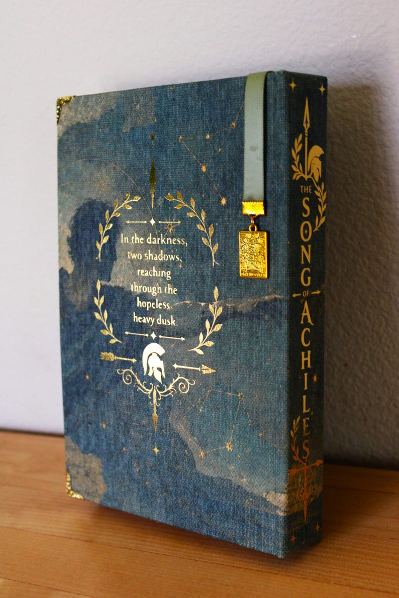 Op de afbeelding: Een blauw gebonden boek getiteld The Song of Achilles met gouden sterrenbeelden en een lauwerkransontwerp. De tekst luidt: In the darkness, two shadows, reaching through the hopeless heavy dusk. Het bevat een groen lint als bladwijzer met een gouden bedel.