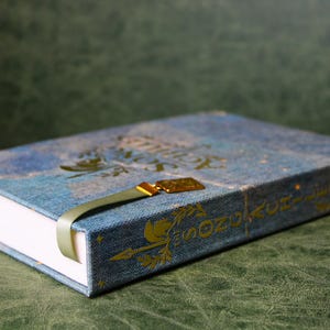 Op de afbeelding: Een blauw hardcover boek met een textiel omslag met gouden letters en decoratieve pijlmotieven. Het boek bevat een lichtgroene lintbladwijzer met een gouden metalen bedel bevestigd aan de bovenkant van de rug van het boek.