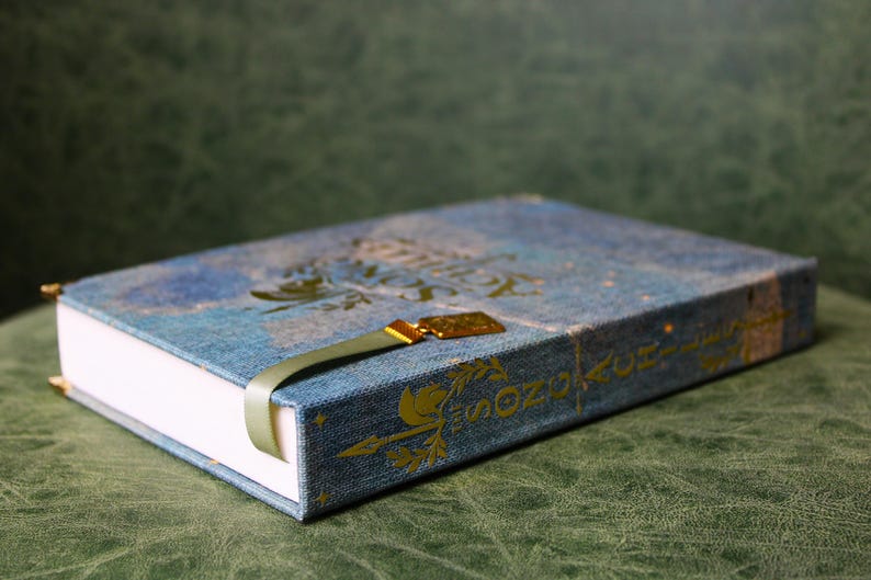 Op de afbeelding: Een blauw hardcover boek met een textiel omslag met gouden letters en decoratieve pijlmotieven. Het boek bevat een lichtgroene lintbladwijzer met een gouden metalen bedel bevestigd aan de bovenkant van de rug van het boek.