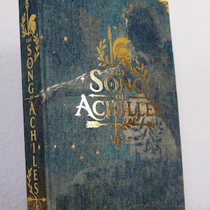 Op de afbeelding: Een hardcover boek getiteld The Song of Achilles van Madeline Miller met een blauwe stoffen kaft met textuur. De voorzijde en rug bevatten goudfolie letters, een helm, een speer en stermotieven. Gouden metalen hoekbeschermers op de kaft.