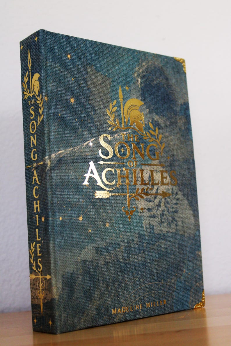 Op de afbeelding: Een hardcover boek getiteld The Song of Achilles van Madeline Miller met een blauwe stoffen kaft met textuur. De voorzijde en rug bevatten goudfolie letters, een helm, een speer en stermotieven. Gouden metalen hoekbeschermers op de kaft.