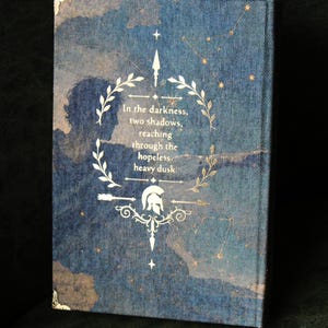 Op de afbeelding: Een hardcover boek met een donkerblauwe stoffen kaft met een sterrenkaartontwerp. De voorkant toont een gouden lauwerkrans, een Spartaanse helm en de tekst: In the darkness, two shadows, reaching through the hopeless, heavy dusk.