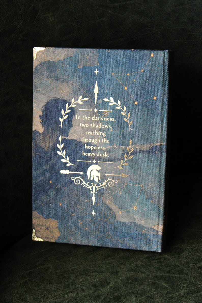 Op de afbeelding: Een hardcover boek met een donkerblauwe stoffen kaft met een sterrenkaartontwerp. De voorkant toont een gouden lauwerkrans, een Spartaanse helm en de tekst: In the darkness, two shadows, reaching through the hopeless, heavy dusk.