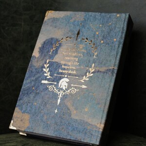 Op de afbeelding: Een hardcover boek met een blauwe textuurkaft voorzien van een goudfolie ontwerp van een helm, lauriertakken en sterren. De tekst luidt: In the darkness, two shadows, reaching through the hopeless, heavy dusk. De hoeken hebben gouden accenten.