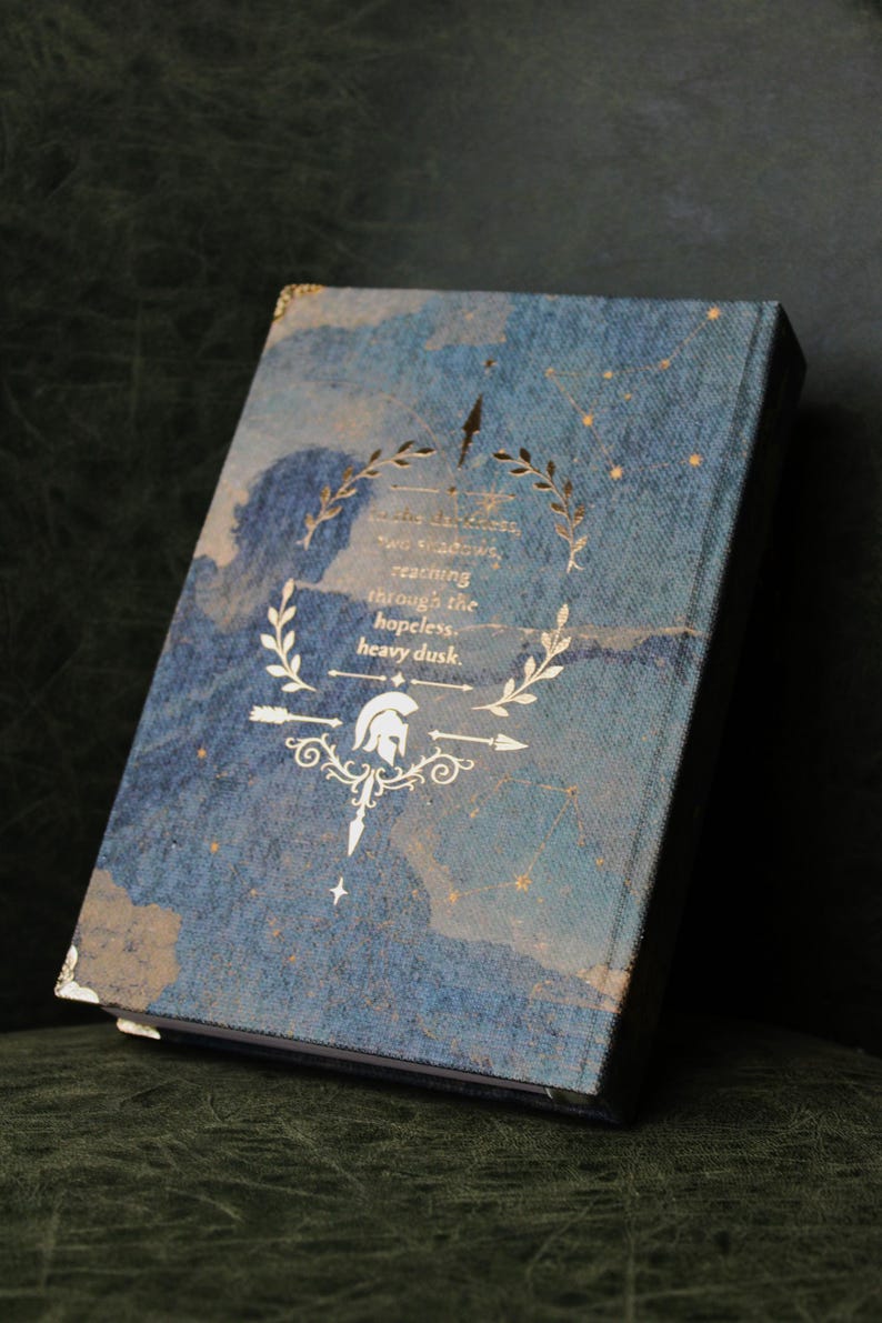 Op de afbeelding: Een hardcover boek met een blauwe textuurkaft voorzien van een goudfolie ontwerp van een helm, lauriertakken en sterren. De tekst luidt: In the darkness, two shadows, reaching through the hopeless, heavy dusk. De hoeken hebben gouden accenten.