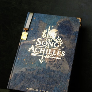 Op de afbeelding: Een hardcover boek getiteld The Song of Achilles door Madeline Miller met een blauwe stoffen kaft, gouden belettering, een helm- en pijlafbeelding, decoratieve hoekaccenten en een lintbladwijzer met een kleine gouden rechthoekige bedel.