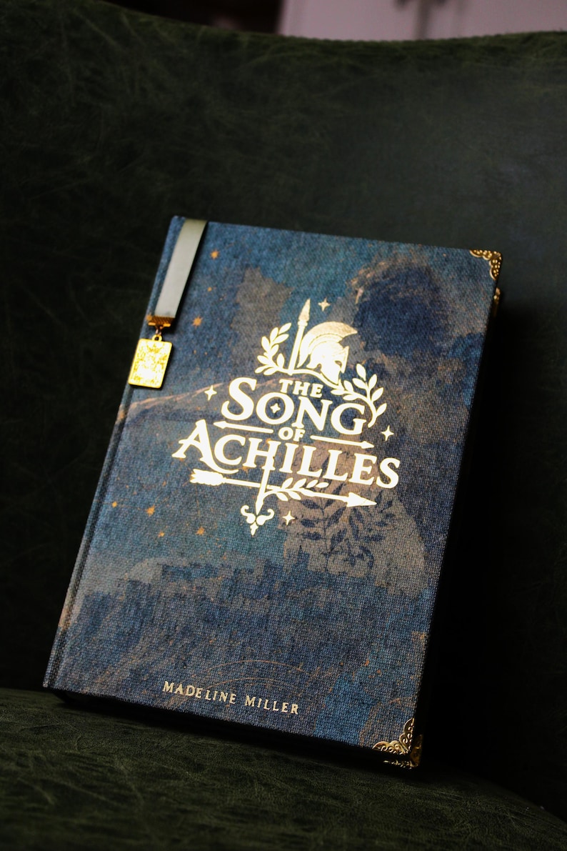 Op de afbeelding: Een hardcover boek getiteld The Song of Achilles door Madeline Miller met een blauwe stoffen kaft, gouden belettering, een helm- en pijlafbeelding, decoratieve hoekaccenten en een lintbladwijzer met een kleine gouden rechthoekige bedel.