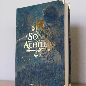 Op de afbeelding: Een hardcover boek getiteld The Song of Achilles door Madeline Miller met een blauwe, gestructureerde stoffen kaft. De voorkant bevat gouden letters, een helm en pijlillustraties. Het boek is voorzien van gouden metalen hoekbeschermers.