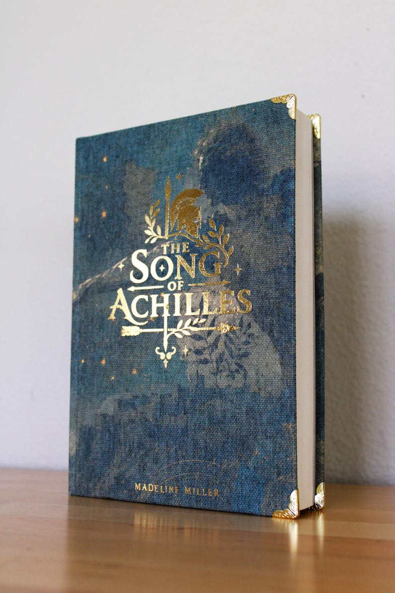 Op de afbeelding: Een hardcover boek getiteld The Song of Achilles door Madeline Miller met een blauwe, gestructureerde stoffen kaft. De voorkant bevat gouden letters, een helm en pijlillustraties. Het boek is voorzien van gouden metalen hoekbeschermers.
