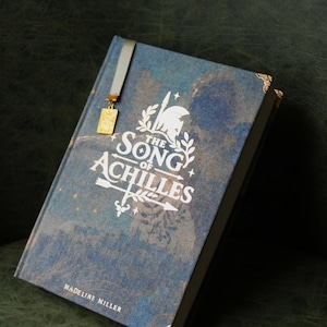 Op de afbeelding: Een hardcover boek getiteld The Song of Achilles door Madeline Miller met een blauwe textuurkaft, witte bedrukte tekst, een afbeelding van een helm en pijl, gouden hoekbeschermers en een grijs lint als bladwijzer met een gouden bedel.