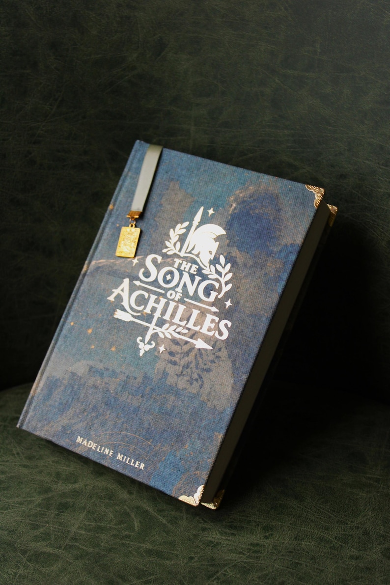 Op de afbeelding: Een hardcover boek getiteld The Song of Achilles door Madeline Miller met een blauwe textuurkaft, witte bedrukte tekst, een afbeelding van een helm en pijl, gouden hoekbeschermers en een grijs lint als bladwijzer met een gouden bedel.