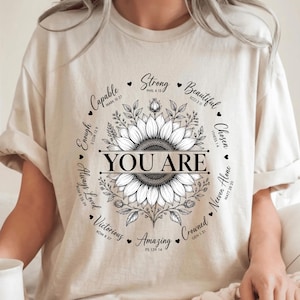 Peut inclure: Un t-shirt beige avec un motif central noir représentant un tournesol entouré du texte "YOU ARE" et de divers mots inspirants tels que "Capable", "Strong", "Beautiful", "Chosen", "Never Alone", "Crowned", "Amazing", "Victorious" et "Always Loved".