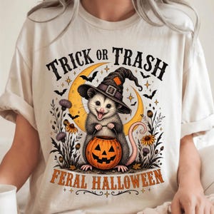 Peut inclure: Un t-shirt couleur crème avec un motif d'opossum portant un chapeau de sorcière et tenant une citrouille d'Halloween. Le texte indique "TRICK OR TRASH" et "FERAL HALLOWEEN" avec un croissant de lune jaune, des chauves-souris et des fleurs.