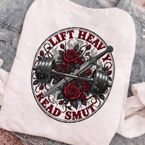 Peut inclure: Un t-shirt blanc avec un motif circulaire représentant une barre de musculation croisée avec une épée, entourée de roses rouges et de vignes épineuses. Le texte LIFT HEAVY READ SMUT est imprimé en rouge sur le bord circulaire du motif.