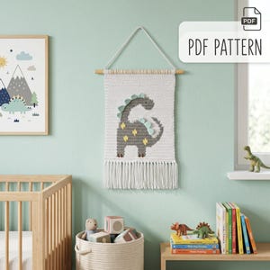 May include: A white crochet wall hanging with a gray dinosaur design, accented with light blue and yellow details. The wall hanging is suspended from a wooden dowel and has white fringe. The text "PDF PATTERN" is in the upper right corner.
