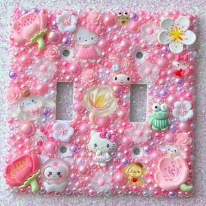 Peut inclure: Un cache-interrupteur rose décoré de perles nacrées, de fleurs et de personnages de dessins animés. Le cache-interrupteur présente Hello Kitty, une grenouille et d'autres motifs mignons.