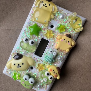 Puede incluir: Una cubierta de interruptor de luz blanca decorada con purpurina verde, estrellas amarillas y varios personajes de dibujos animados. Los personajes incluyen un perro amarillo con una boina marrón, una rana verde y un pollito amarillo.