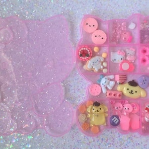 May include: A pink plastic container shaped like a cartoon character with multiple compartments filled with small colorful charms and figurines. The container is open and the charms are visible.