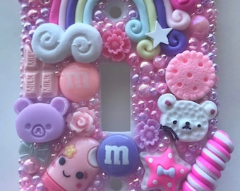 May include: A decorative light switch cover, heavily embellished with pink and purple resin decorations. Features a rainbow, stars, a narwhal, a lollipop, and various other cute, colorful charms. The background is covered in small, iridescent beads.