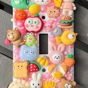 May include: A white light switch cover decorated with colorful, cartoon-style food and animal-shaped embellishments. The cover features a green dinosaur, panda, bear, cheese, apple, bunny, and other cute designs. The background is filled with pink beads.