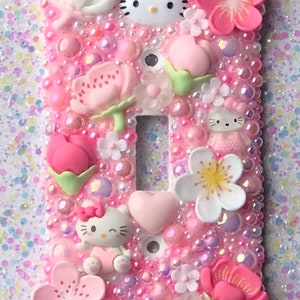 Peut inclure: Un cache-interrupteur blanc décoré de fleurs roses, blanches et jaunes, de cœurs roses et blancs et de personnages Hello Kitty. Le cache est recouvert de paillettes roses.