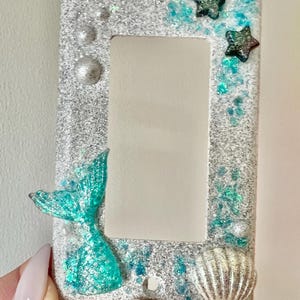 May include: A decorative light switch cover with a mermaid theme. The cover is silver glitter with turquoise accents, including a mermaid tail, starfish, and seashell. The cover has a rectangular opening for the switch.