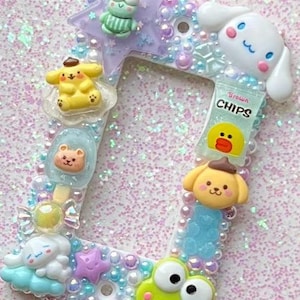 Puede incluir: Un marco de fotos decorativo con borde blanco, adornado con varios personajes de dibujos animados y adornos. El marco presenta una estrella, una rana, un perro, un helado de oso y una bolsa de patatas fritas. El marco está decorado con perlas y purpurina.