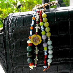 May include: A decorative bag charm featuring three strands of multi-colored beads, including green, red, blue, and clear glass, attached to a gold-toned metal ring. The charm hangs from a black, textured, crocodile-embossed leather handbag.