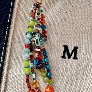 May include: A gold-toned metal keyring featuring a multi-strand beaded tassel. The strands consist of various glass, stone, and plastic beads in shades of red, orange, yellow, green, and blue, accented with a central teal-colored metal seashell charm.