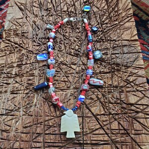 May include: A necklace featuring a central cream-colored bird-shaped pendant suspended from a strand of small red square beads interspersed with various multi-colored glass charms, including blue, white, and iridescent rectangular and round shapes.