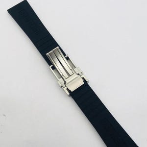May include: A black rubber watch strap featuring a silver-toned metal deployment clasp. The strap has a textured surface with horizontal ridges and the word BREITLING embossed on the upper section of the band in a matte black finish.