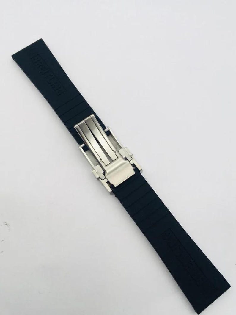 May include: A black rubber watch strap featuring a silver-toned metal deployment clasp. The strap has a textured surface with horizontal ridges and the word BREITLING embossed on the upper section of the band in a matte black finish.