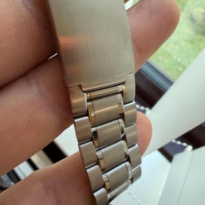 May include: Close-up of a stainless steel watch band clasp. The word "Speedmaster PROFESSIONAL" is engraved on the clasp. The band is made of interlocking metal links.
