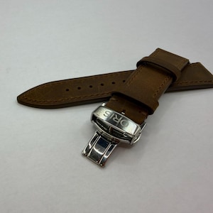 May include: A brown leather watch strap featuring a silver-toned metal deployment clasp. The clasp is engraved with the brand name ORIS. The strap has a matte finish with visible stitching along the edges and multiple adjustment holes.