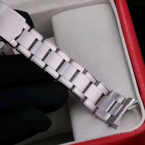 Pu&ograve; includere: Un cinturino per orologio in metallo color argento caratterizzato da una serie di maglie rettangolari lucide collegate da un meccanismo di chiusura sicuro. Il cinturino presenta una superficie liscia e riflettente con perni visibili.