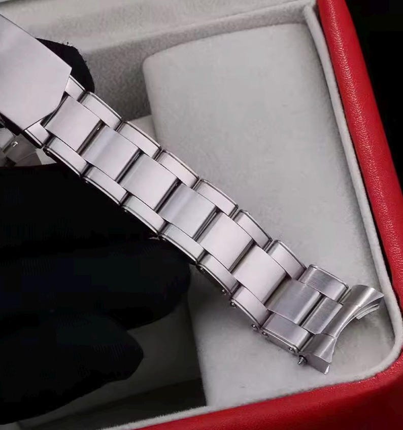 Pu&ograve; includere: Un cinturino per orologio in metallo color argento caratterizzato da una serie di maglie rettangolari lucide collegate da un meccanismo di chiusura sicuro. Il cinturino presenta una superficie liscia e riflettente con perni visibili.