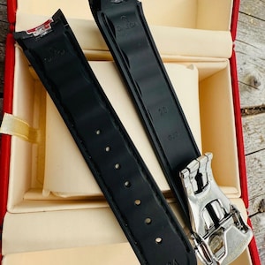 May include: Black watch straps with the Omega logo and a silver clasp are displayed inside a red box. The straps have the numbers 20 and 64 printed on them.