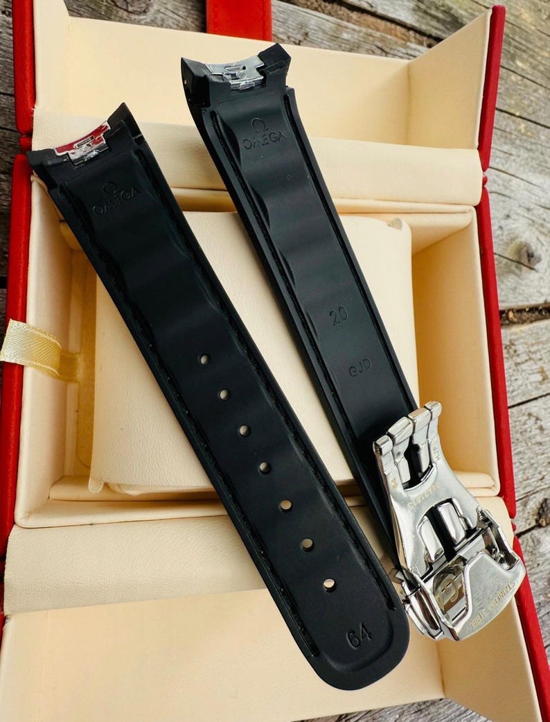 May include: Black watch straps with the Omega logo and a silver clasp are displayed inside a red box. The straps have the numbers 20 and 64 printed on them.