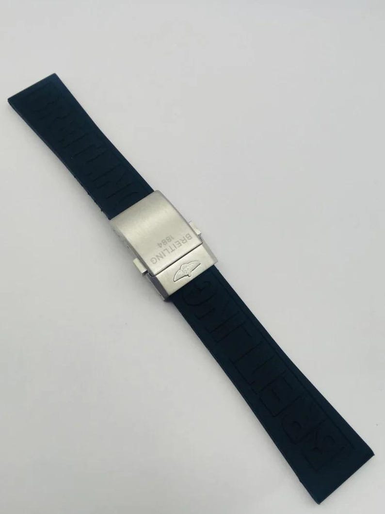 May include: A black rubber watch strap featuring the embossed word BREITLING along the band. The strap includes a brushed silver-tone metal deployment clasp engraved with the text BREITLING 1884 and a small winged logo emblem on the surface.
