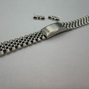 May include: A silver-toned metal watch bracelet featuring a multi-link jubilee design with a brushed finish folding clasp. The clasp is engraved with the word SEIKO. Two additional loose metal end links are positioned above the main bracelet.