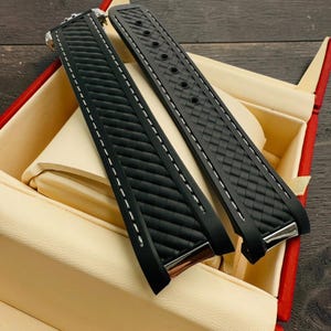 May include: Two black watch straps with white stitching and silver-tone hardware are displayed inside a red and cream-colored box. The straps have a textured pattern and are made of a flexible material.